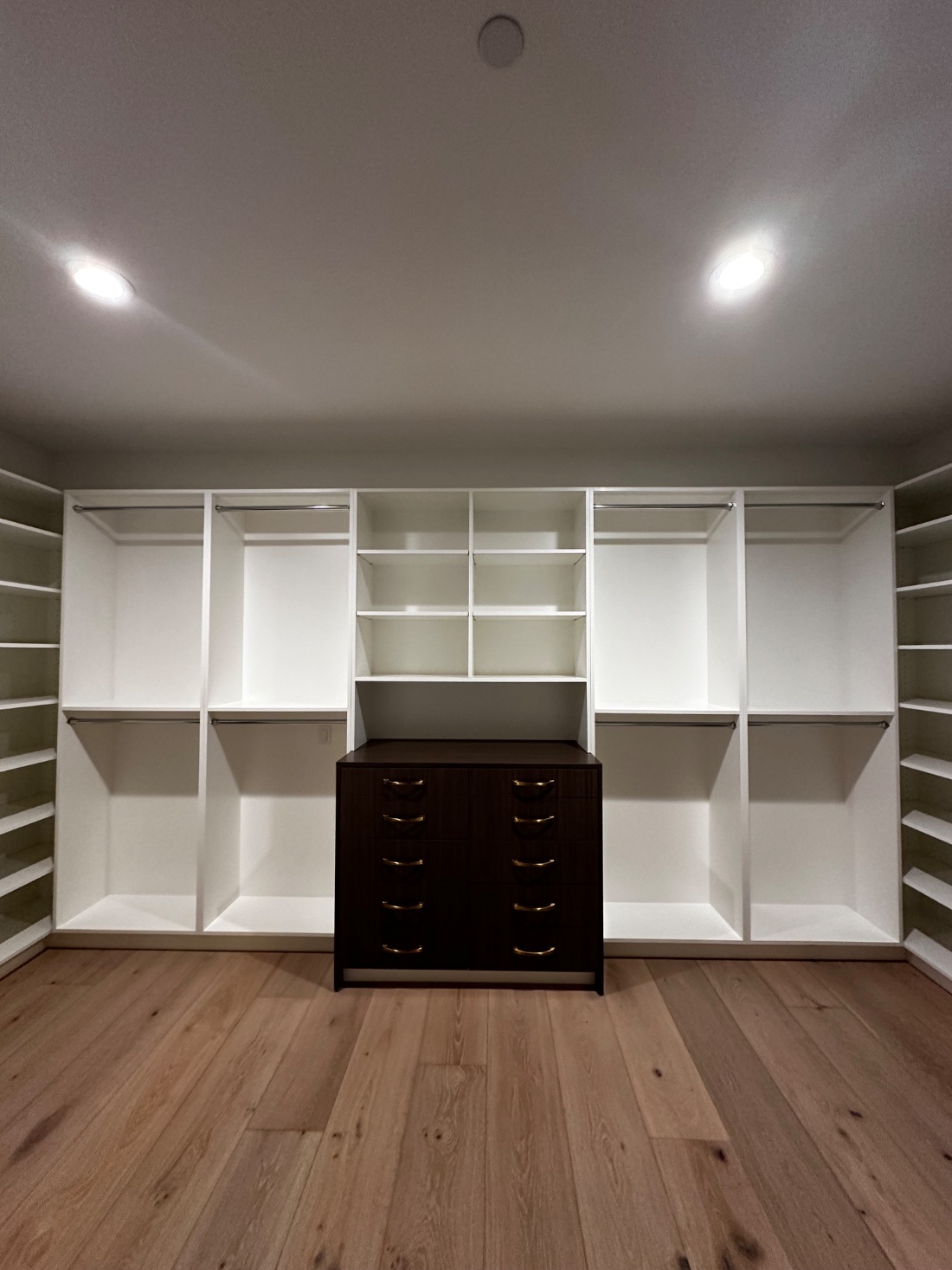 A walk-in closet with white shelving, brass hanging rods, and a dark wood drawer tower at center.